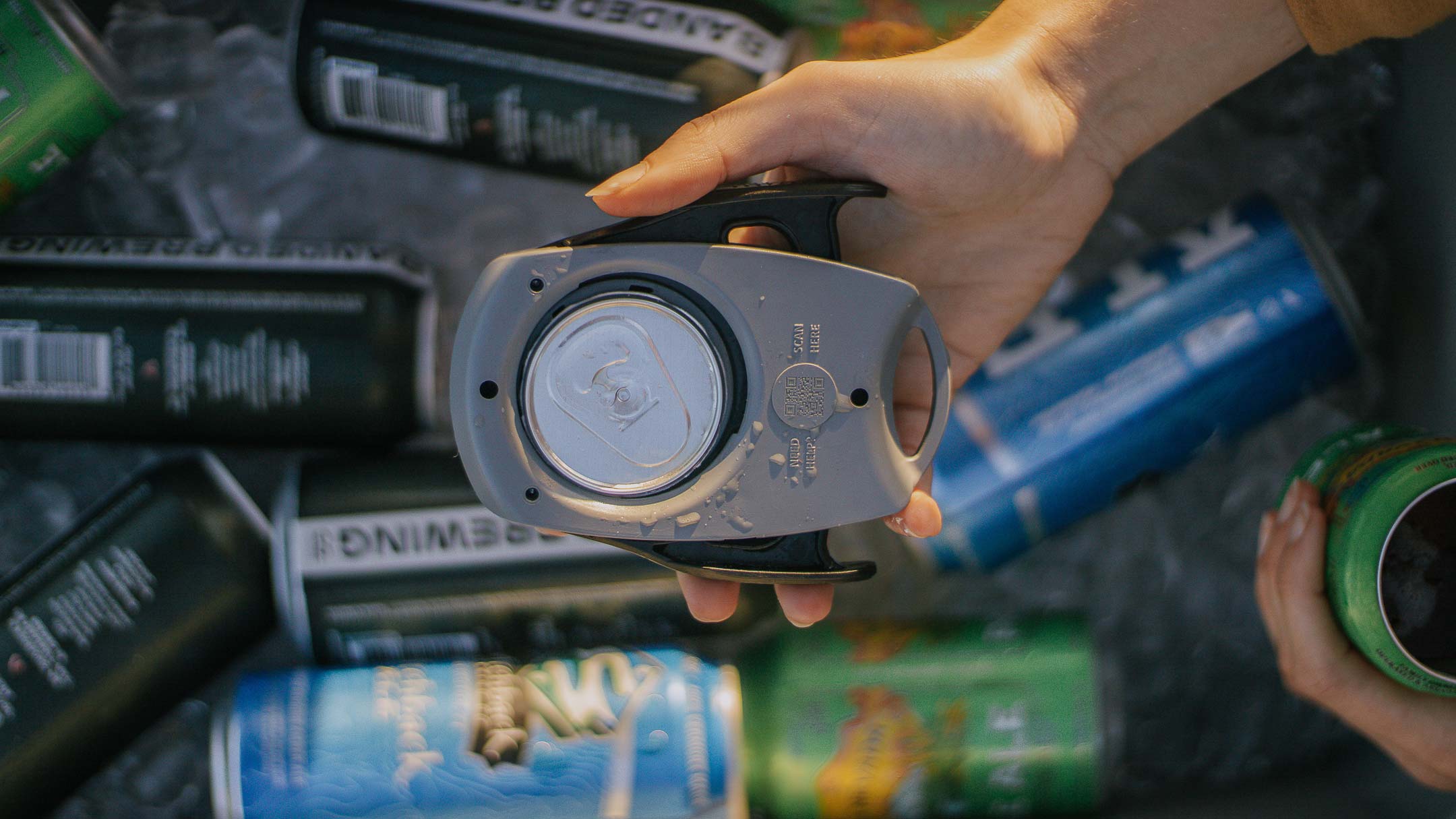 A person holds a can opener for removing the entire top of a beverage can, with various cans and ice visible in the background.